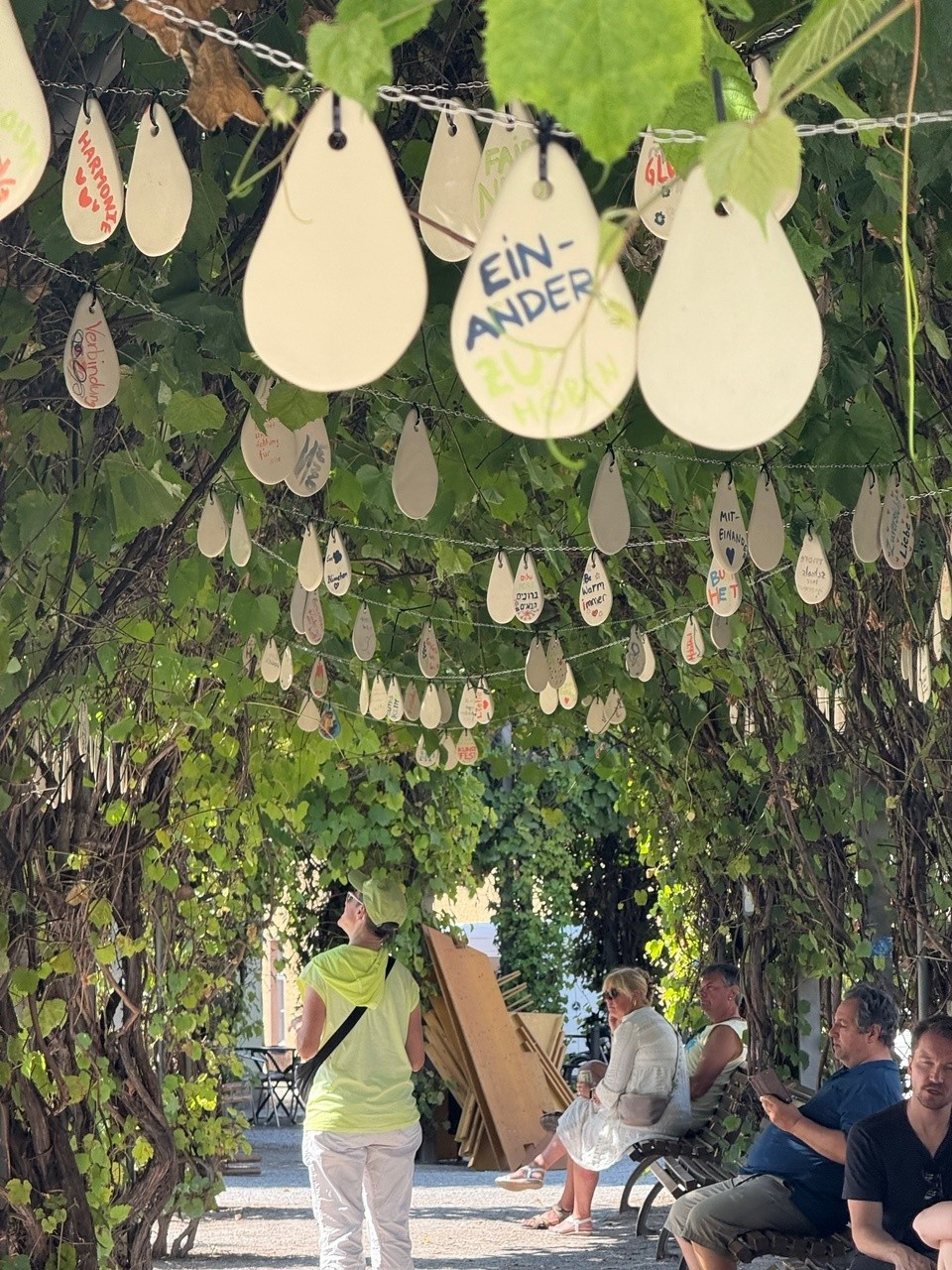 Teardrop-shaped ceramic tiles hang in a garland between trees, beneath which people walk.