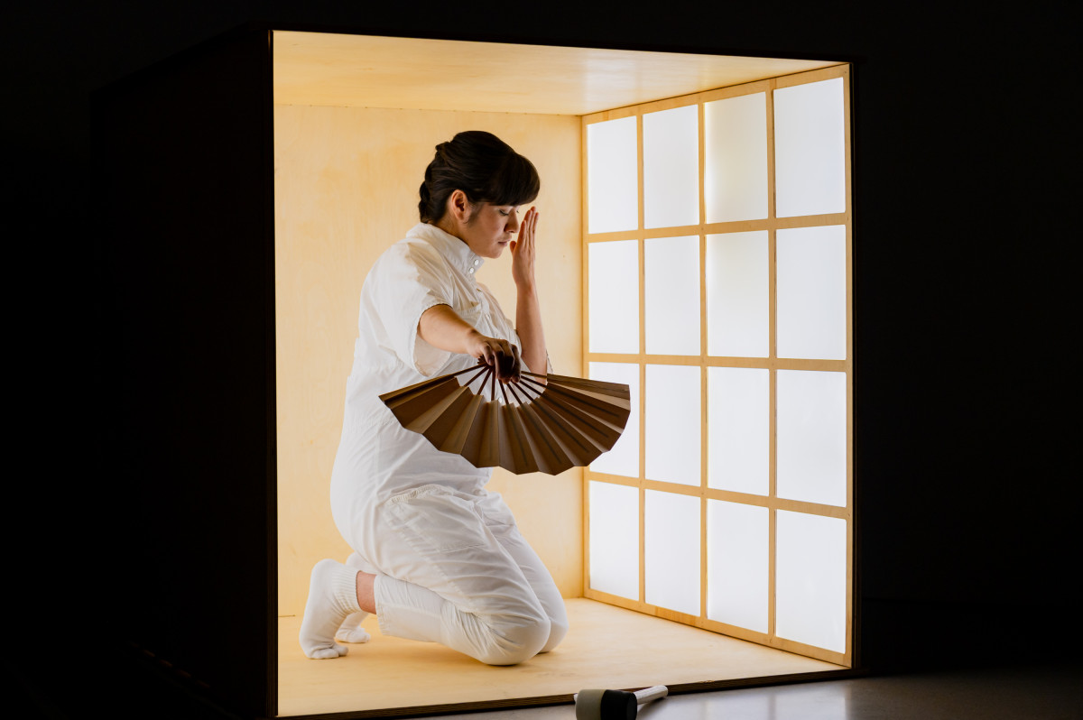 A woman in a brightly illuminated box holds a fan out in front of her. She holds her other hand over her nose. She is dressed in white and, kneeling, just about fits into the box.