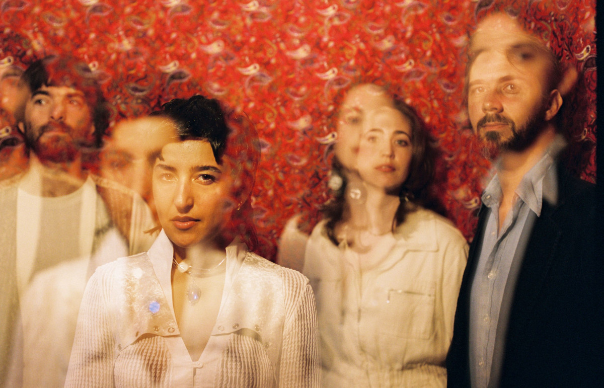 Four people are standing in front of a wall with a red pattern. The two women are wearing white clothes. Of the men, one is wearing white clothes and the other is wearing black and blue. All four people appear as double images overlapping each other.