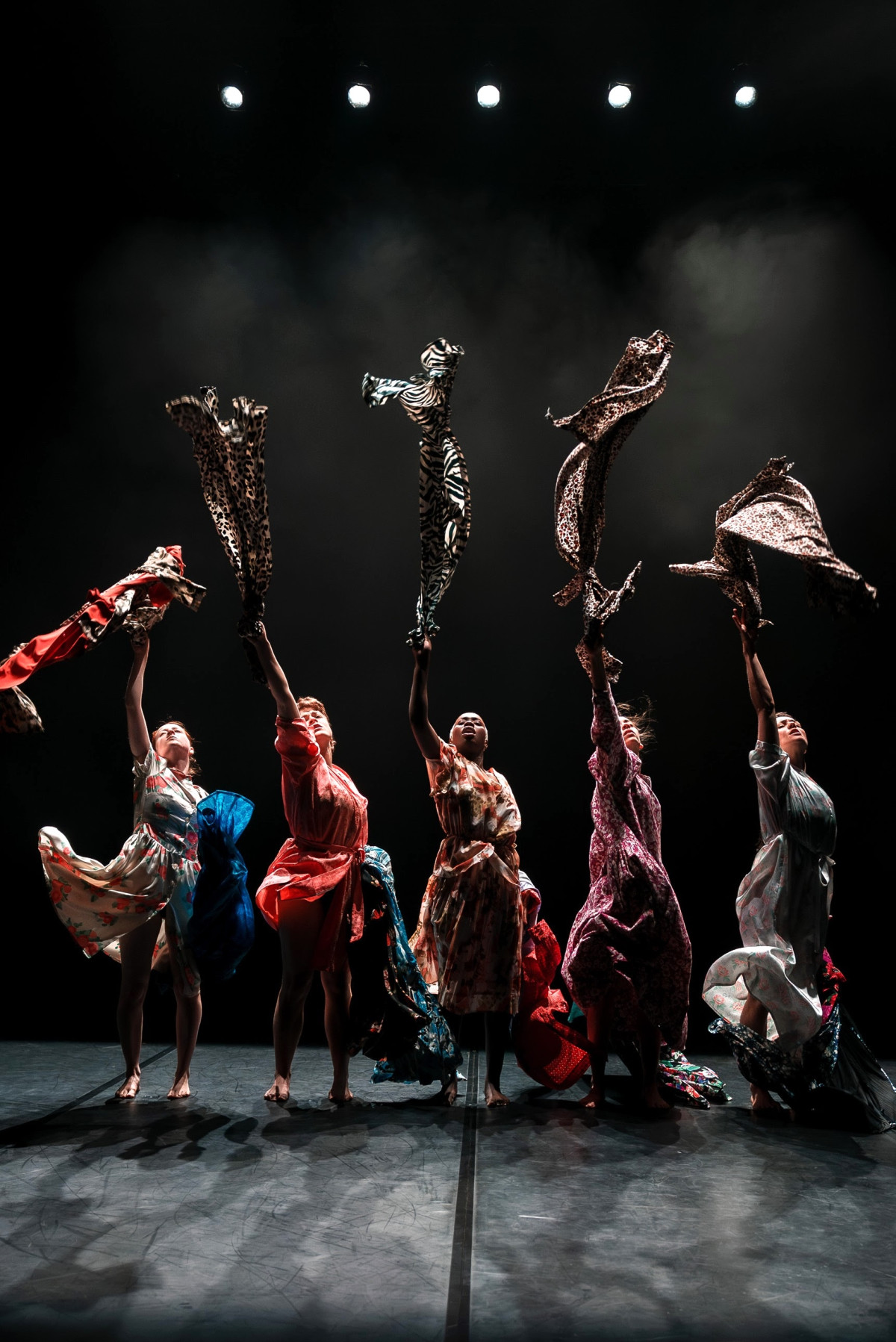 Five women are standing in a row wearing colourful dresses. They are holding up a clothing piece with their right arms. The background is dark. They are barefoot.