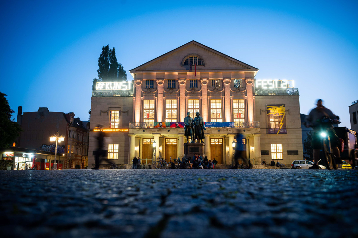 Theatre Square, the Goethe-Schiller monument and the entrance to the DNT are all lit up at dusk. On the balcony of the DNT on the left it says: “#Art”, and on the balcony on the right it says: “Fest 21”.