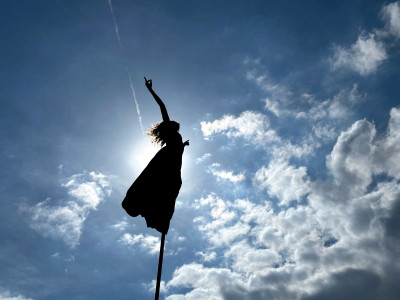 Eine Person mit Rock ist auf einer Stange im Himmel. Die Person hat lange Haare und verdeckt die Sonne. Der Himmel ist Blau.