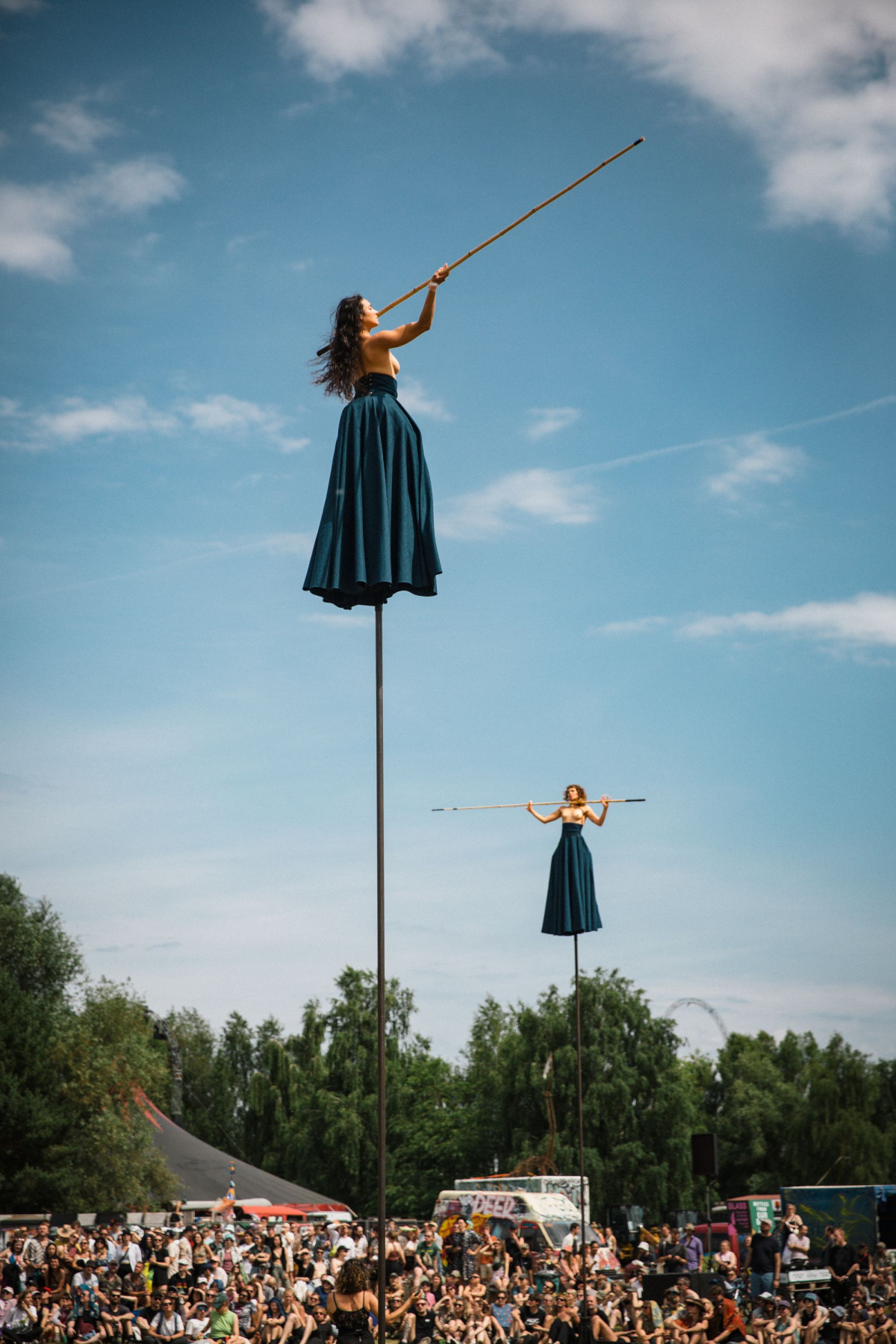 Two people wearing long blue skirts are standing on long sticks while many people watch from the ground. The people are bare-chested and are holding sticks in their hands.
