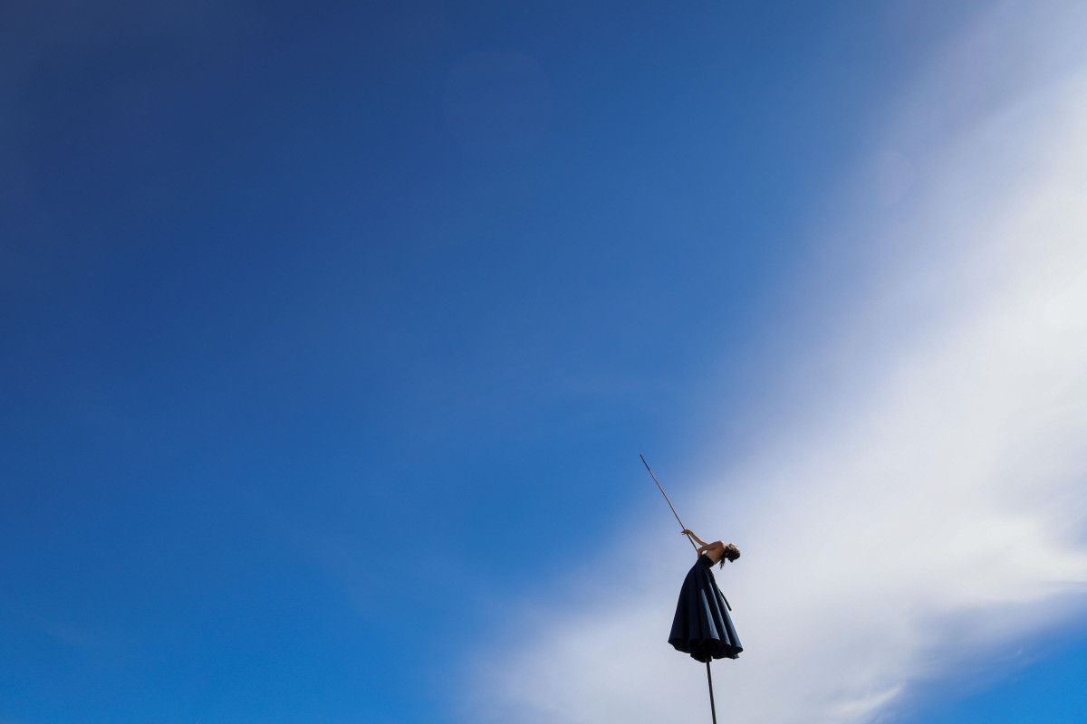 A person in a skirt is standing on a pole in the sky, holding a pole in their hand. The skirt and the sky are blue.