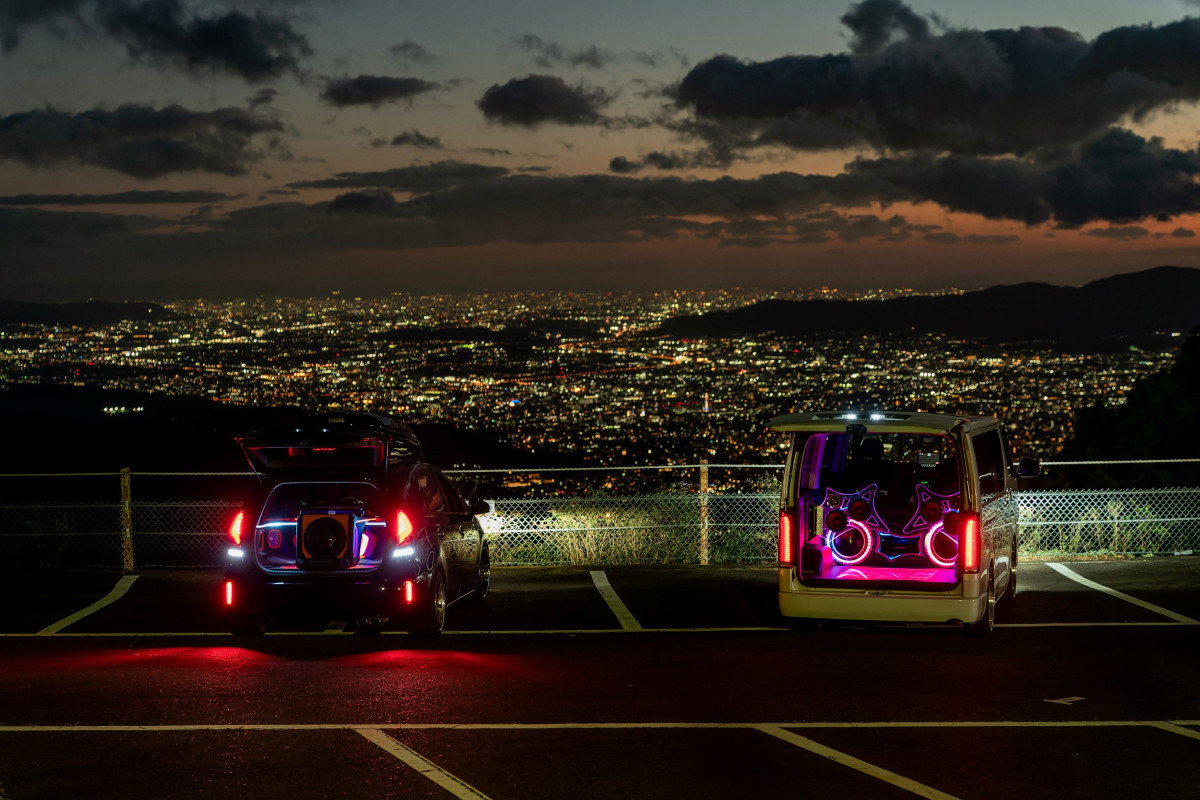 Two cars with open trunks containing jukeboxes and lighting are parked in front of a city lit up at dusk. The cars are in a parking lot on a hill. They look down on the big city. The jukeboxes glow pink and take up half the trunk.