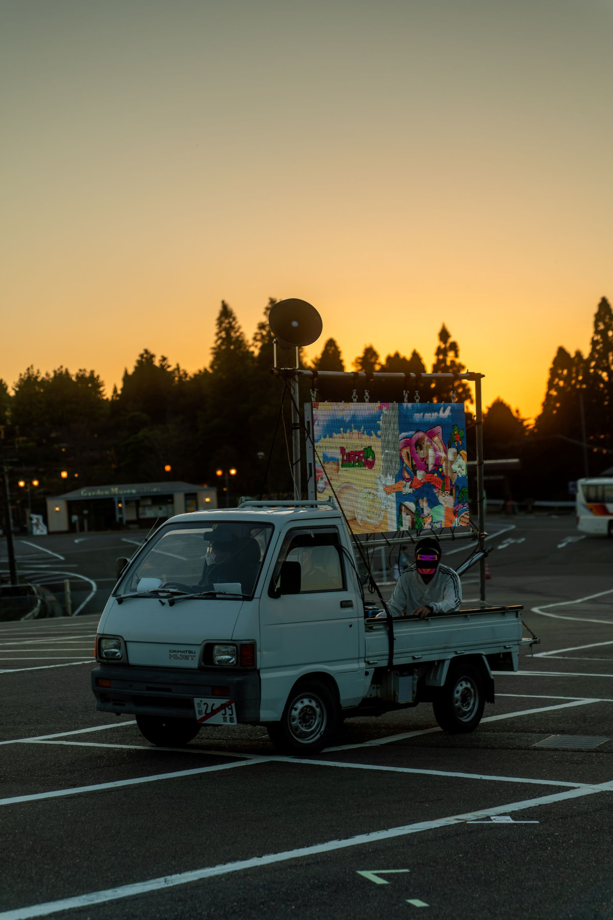 A truck carrying a person and a screen drives across a car parking lot at sunset. The person on the flatbed trailer of the white truck is wearing an LED mask that lights up. The screen above them displays colourful cartoons. A megaphone is attached to the screen. The car parking lot is empty.
